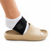 A beige open-toe slide sandal with a black ankle support strap worn around a person's ankle, shown against a plain white background.